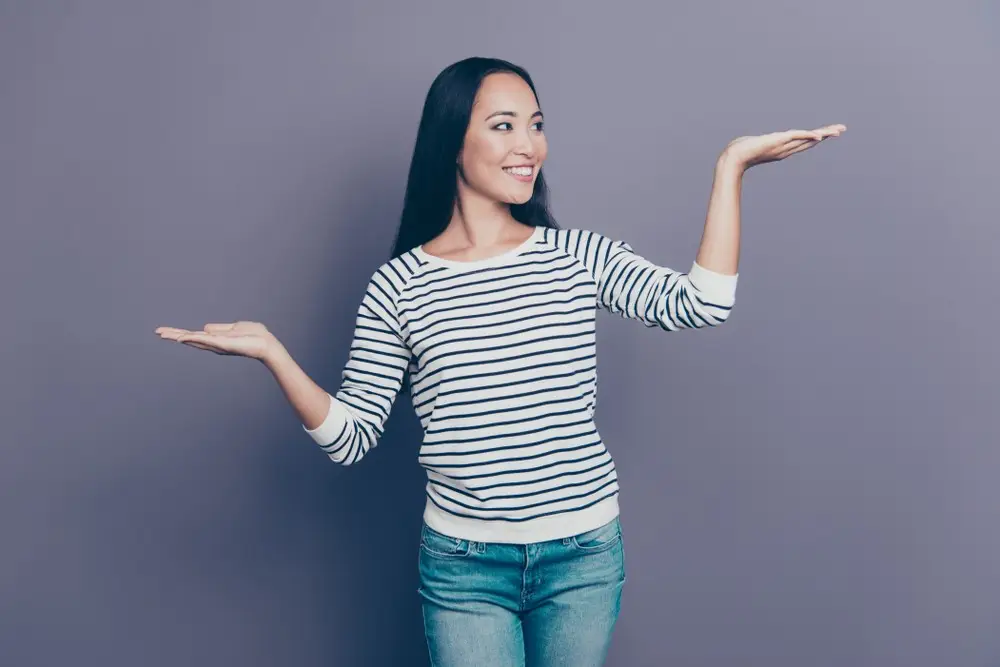A woman in a striped long-sleeve shirt and jeans stands against a plain purple background, smiling and holding her hands up, as if presenting options like liposuction Dallas or a tummy tuck.