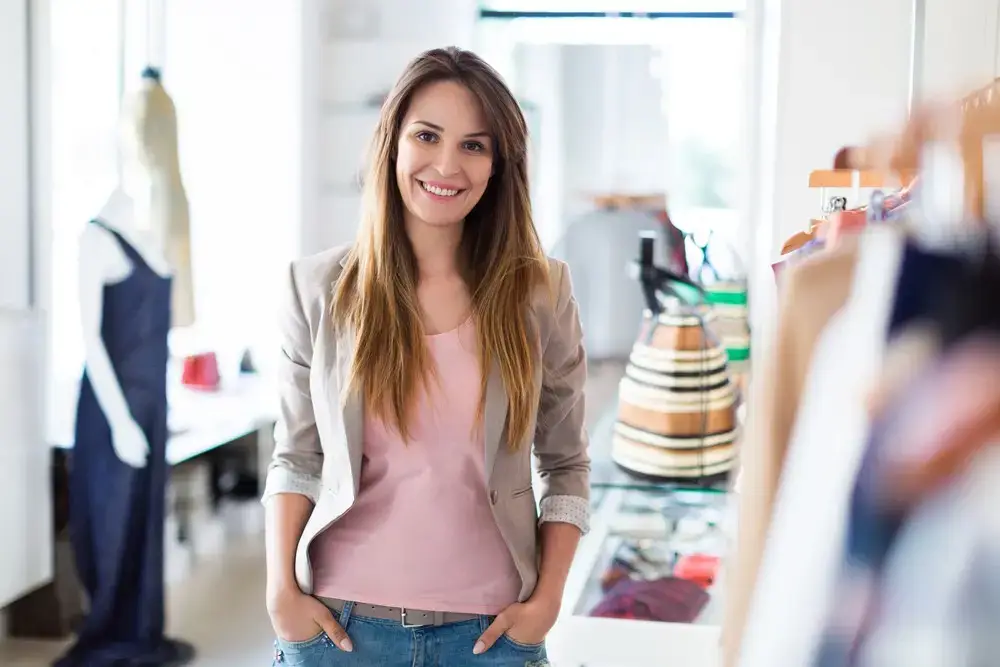 A woman with long brown hair, wearing a beige blazer and pink shirt, stands in a bright boutique with hands in her pockets, smiling at the camera. Clothing racks and a mannequin hint at her passion for body sculpting and style.