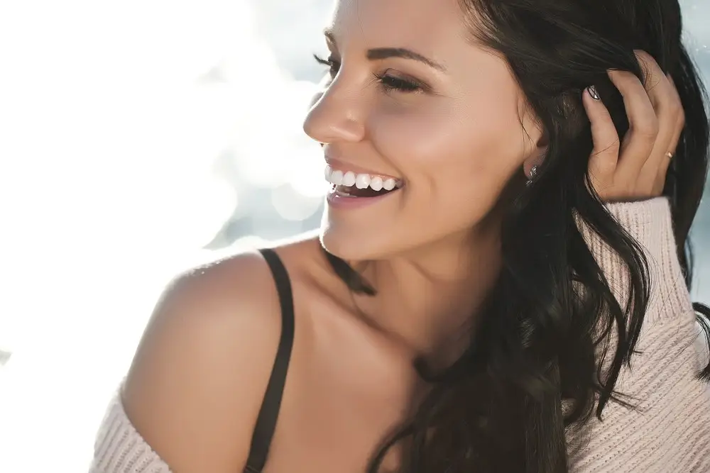 A woman with long dark hair smiles while looking to the side, her hand touching her hair. She wears a light off-the-shoulder sweater, revealing a black strap. The bright, blurred background highlights her confidence after body sculpting.
