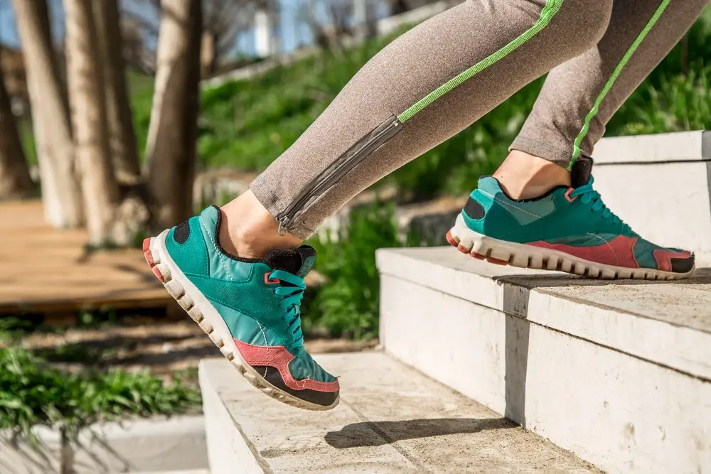 A person wearing teal and pink athletic shoes and light brown leggings with a green stripe is running or stepping up concrete stairs outdoors, perhaps showcasing the benefits of body sculpting, with greenery and trees in the background.