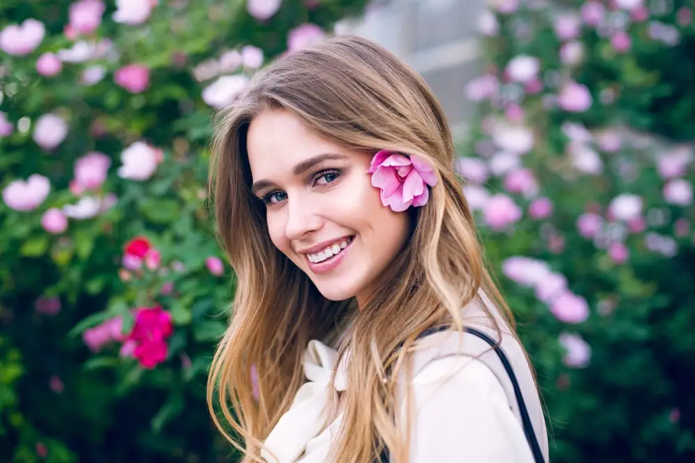 A young woman with long, light brown hair smiles at the camera, standing outdoors with pink flowers in the background and a pink flower tucked behind her ear, radiating confidence after her tummy tuck.