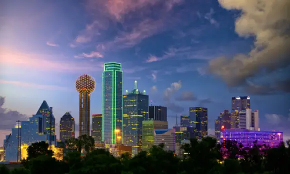 Downtown Dallas skyline at dusk, featuring illuminated skyscrapers with colorful lights—including the green-outlined Bank of America Plaza and round Reunion Tower—creates a stunning backdrop for those seeking body sculpting or a tummy tuck in the city.