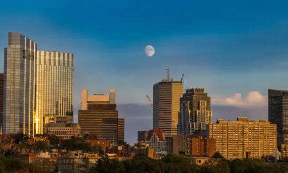 A city skyline with modern high-rise buildings and a half moon visible in a blue sky during daylight. The buildings reflect sunlight, greenery is visible in the foreground—much like the fresh confidence after a liposuction Dallas procedure.