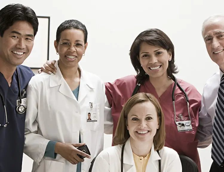 Five healthcare professionals, three women and two men, smile at the camera in a clinical setting. Dressed in lab coats and scrubs with stethoscopes, they specialize in procedures like body sculpting and fat grafting.