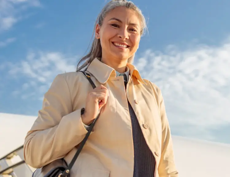 A person with gray hair in a ponytail, wearing a beige coat and carrying a black shoulder bag, smiles while standing outdoors under a blue sky with scattered clouds, embodying confidence often associated with body sculpting.