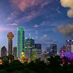 A city skyline at dusk featuring tall buildings with green and purple lights, the sky tinged with pink and blue clouds, and trees silhouetted in the foreground—much like the vibrant possibilities of cosmetic surgery Texas offers.